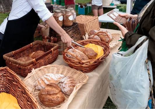 A farmers market table with woven baskets of fresh sourdough bread, a vendor in an apron, and a customer shopping